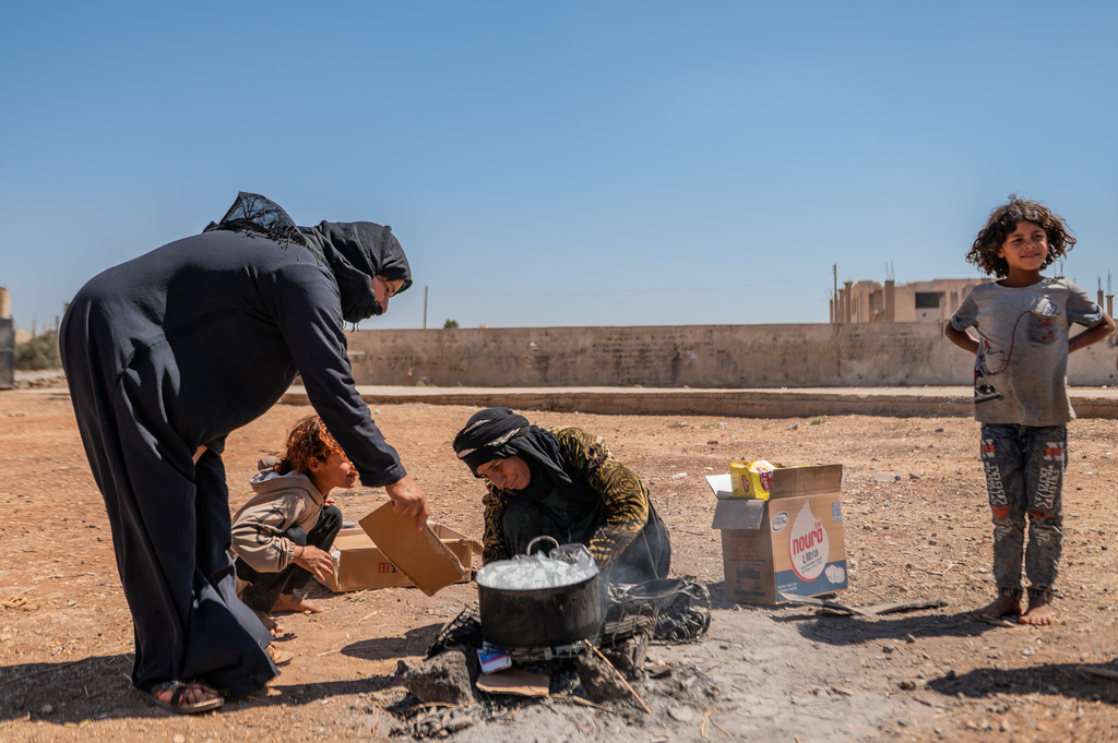 Image of two women and one girl around a pot of food on a sunny day in an enclosure