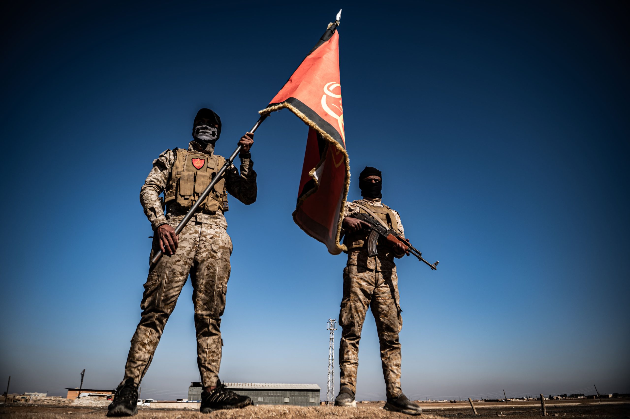 Two men in combat fatigues, one of them holding a flag, faces covered